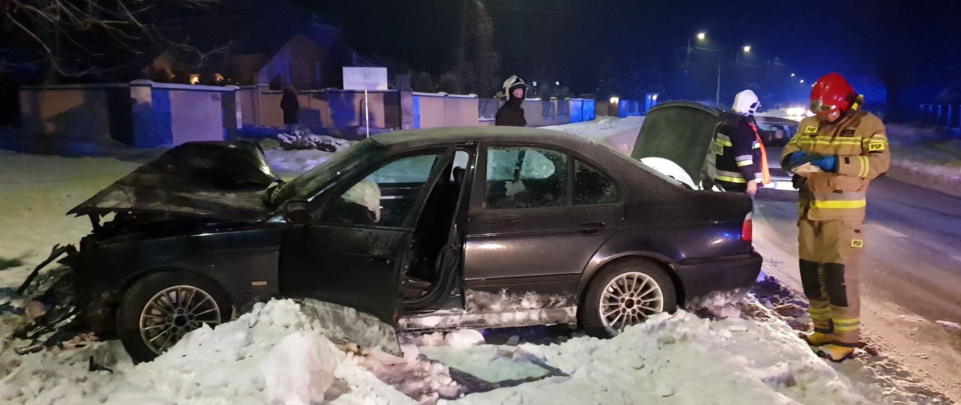Na zdjęciu widać uszkodzony pojazd marki BMW który znajduje się na poboczu drogi. Trzech ratowników straży pożarnej usuwa skutki zdarzenia. 