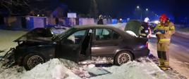 Na zdjęciu widać uszkodzony pojazd marki BMW który znajduje się na poboczu drogi. Trzech ratowników straży pożarnej usuwa skutki zdarzenia. 