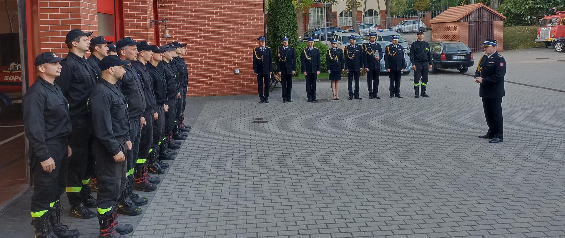 stażacy po lewej stronie stoją w dwuszeregu , na wprost funkcjonariusze biurowi a po prawej komendant przemawia