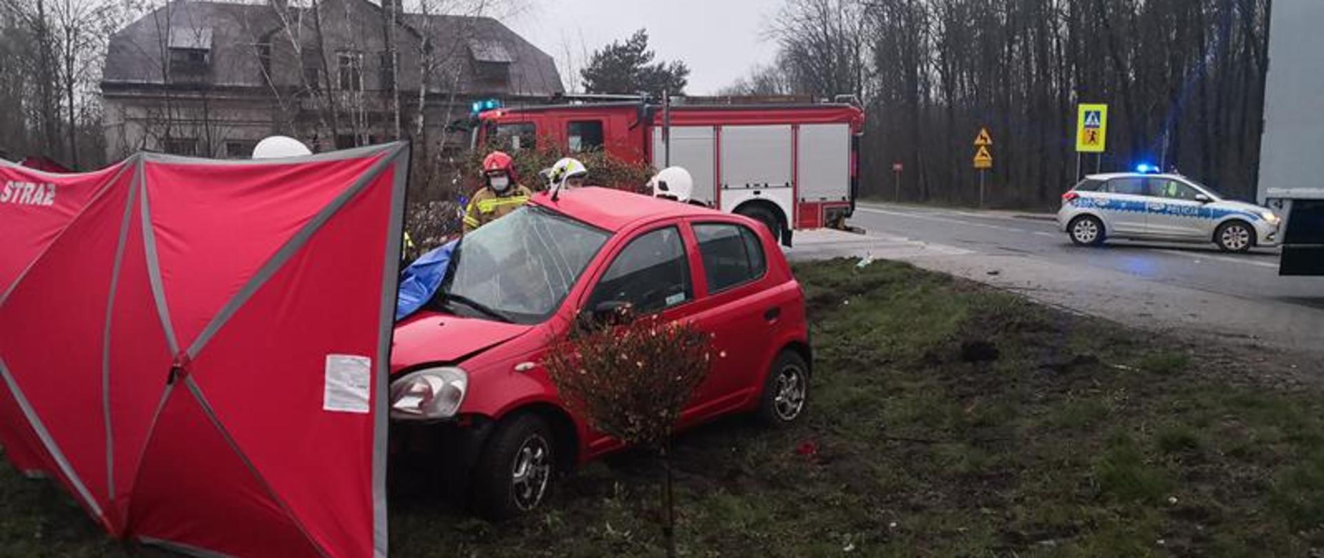 Uszkodzony samochód który uczestniczył w zdarzeniu znajduje się poza drogą na pasie zieleni. Czerwony parawan zasłania działania udzielania pomocy przez ratowników osobie poszkodowanej