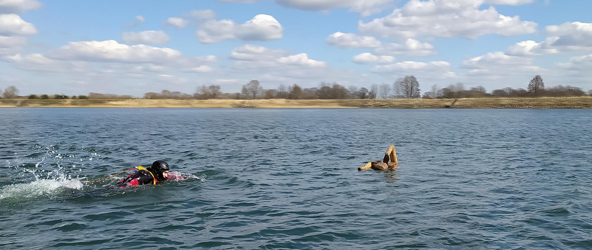 Strażak ubrany w suchy skafander płynie do drewnianego manekina, który pozoruje osobę poszkodowaną.