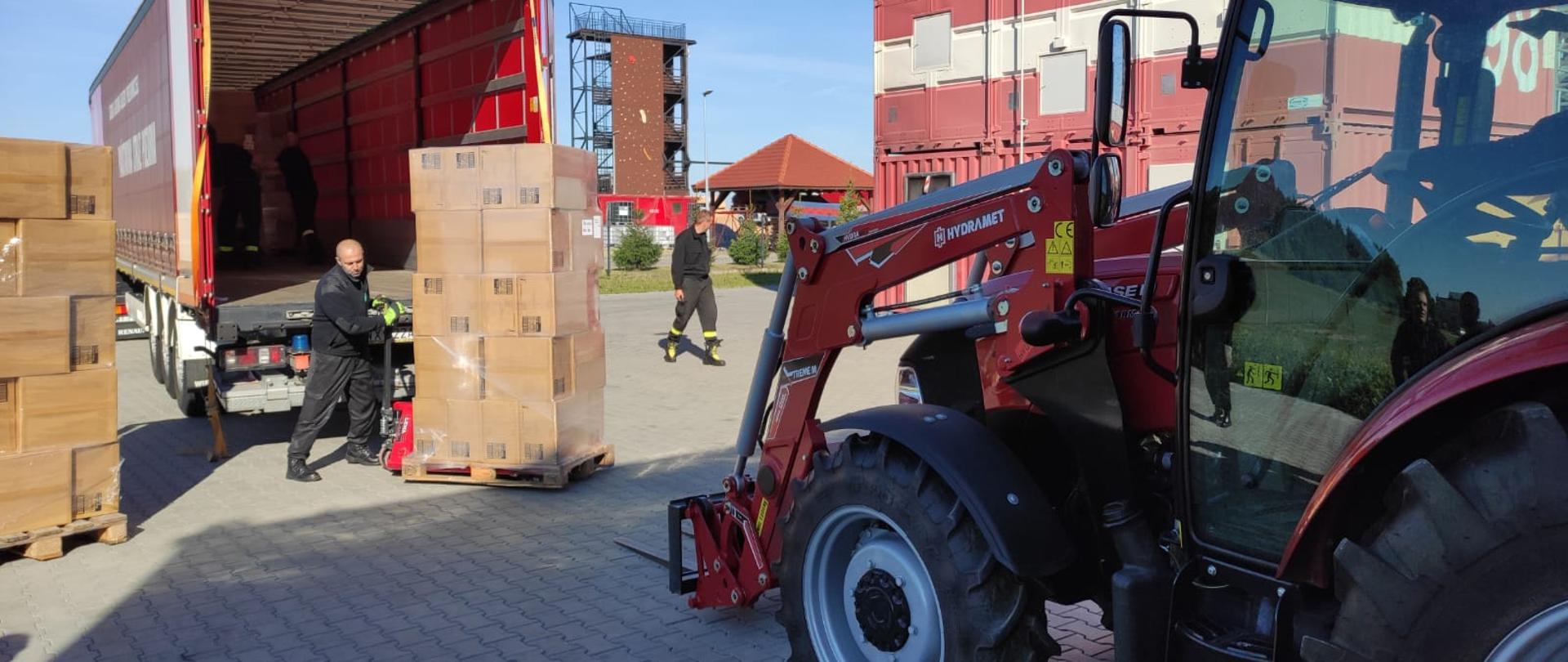 Widok na załadunek naczepy kontenera. Na pierwszym planie ciagnik z ładowaczem, tuz przed nim palety ze sprzętem, zapakowane. Przy paletach strażak. W tle obiekty ośrodka szkolenia.