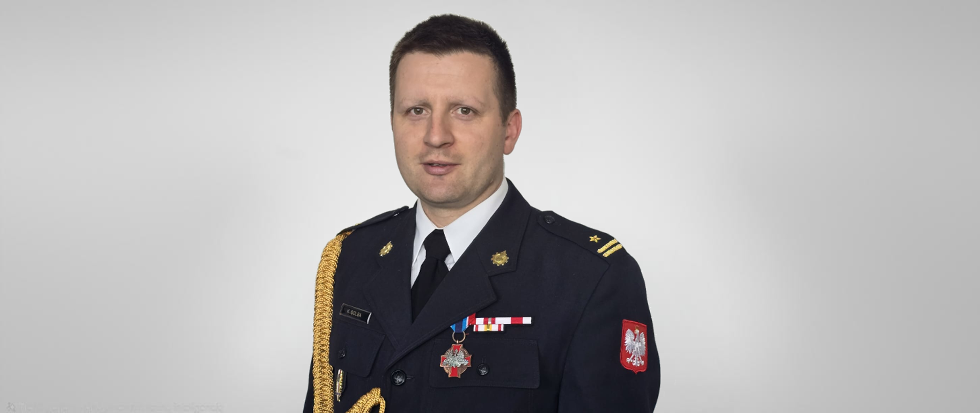 Komendant PSP na zdjęciu w mundurze wyjściowym. Zdjęcie obejmuje popiersie Komendanta. Na mundurze widoczne odznaczenia pożarnicze oraz imiennik