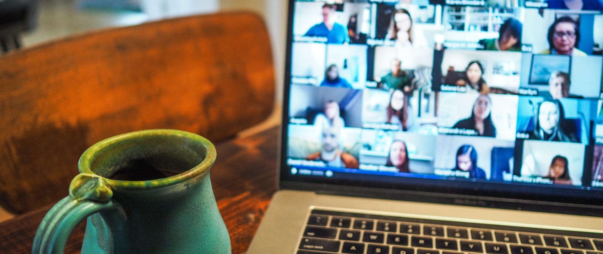 Zdjęcie poglądowe: komputer na biurku. Na komputerze widać wiele ekranów z uczestnikami spotkania. Obok komputera kubek