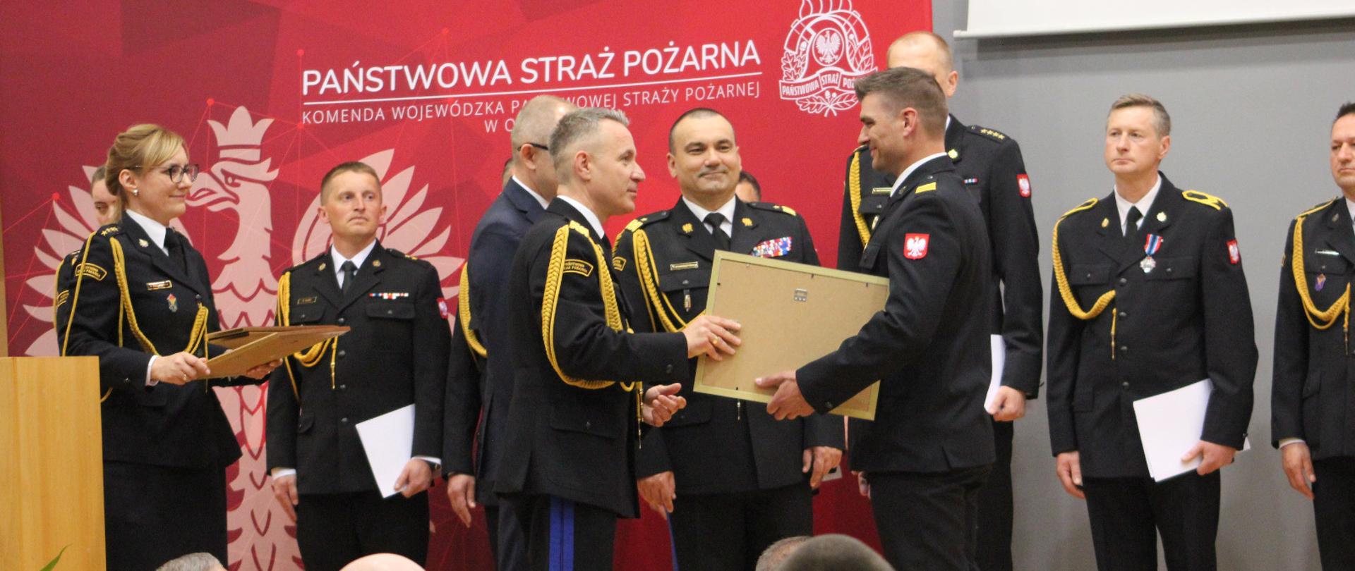 Komendant główny PSP wręczający akt mianowania dla strażaka PSP. W tle strażacy PSP 