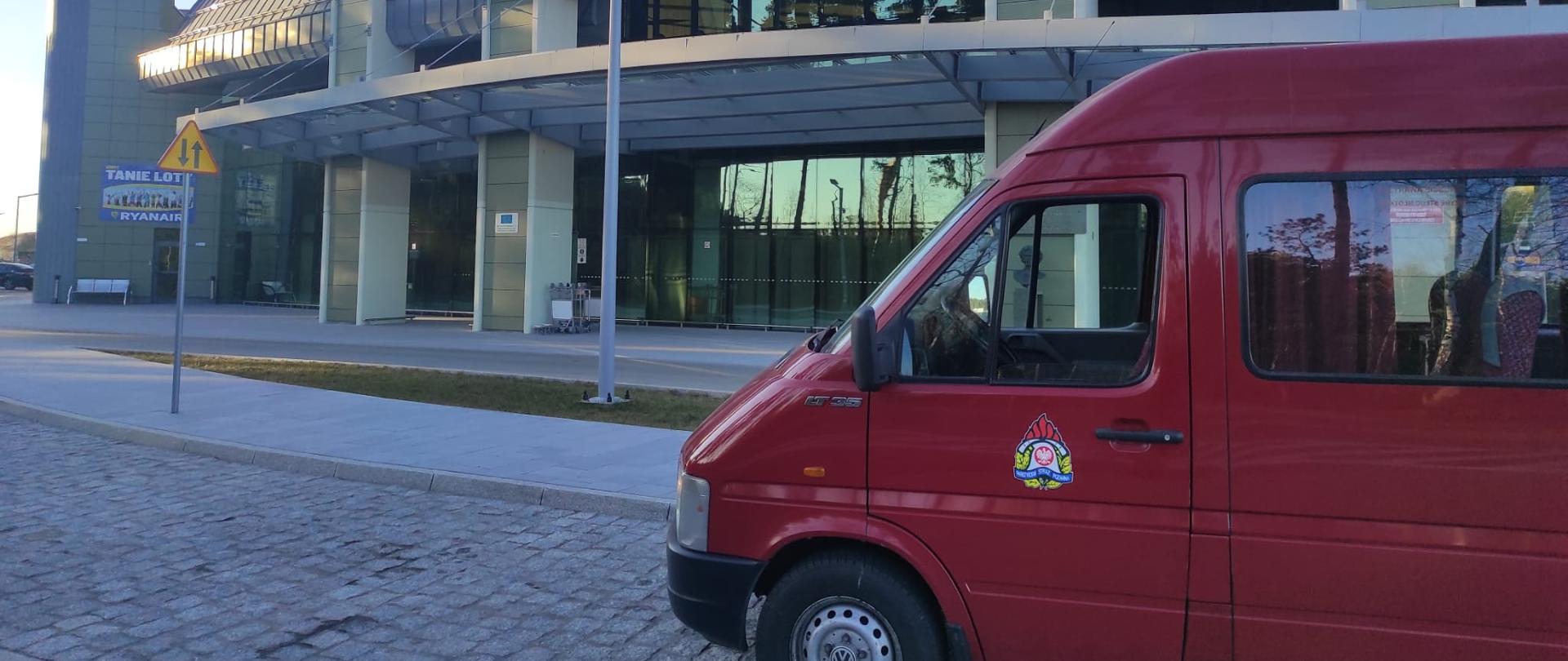 na pierwszym planie z lewej strony widać prawą przednią stronę busa czerwonego z herbem PSP na drzwiach kierowcy. W tle budynek dworca lotniczego w Bydgoszczy. Szara elewacja, w środkowej części budynku szary napis Bydgoszcz.