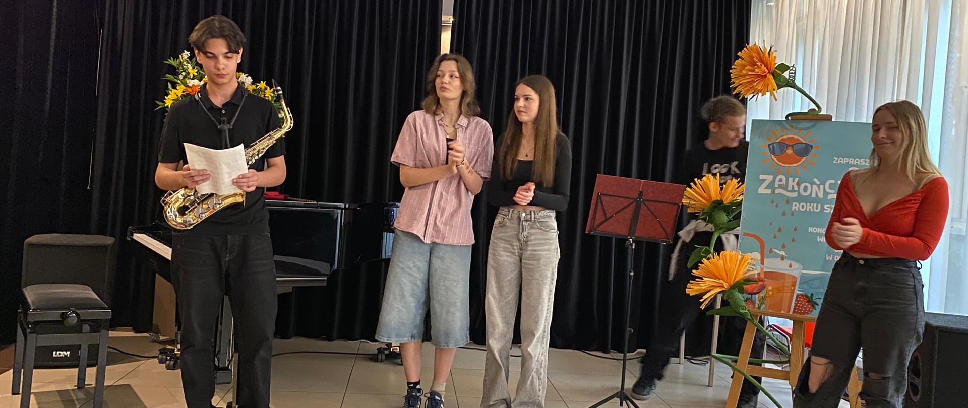 Na scenie stoi kilkoro wychowanków. Po lewej stronie przed fortepianem stoi chłopiec z mikrofonem, pośrodku dwie dziewczyny klaszczące z dłonie. Z prawej strony na sztaludze stoi plakat informujący o zakończeniu roku. Na plakacie na niebieskim tle na górze jest słońce w okularach przeciwsłonecznych, a na dole napój w szklance ze słomką i truskawka. Po lewej stronie plakatu widać chłopca, trzy duże kwiaty z bibuły i pulpit, a z prawej stoi dziewczyna w czerwonej bluzce.