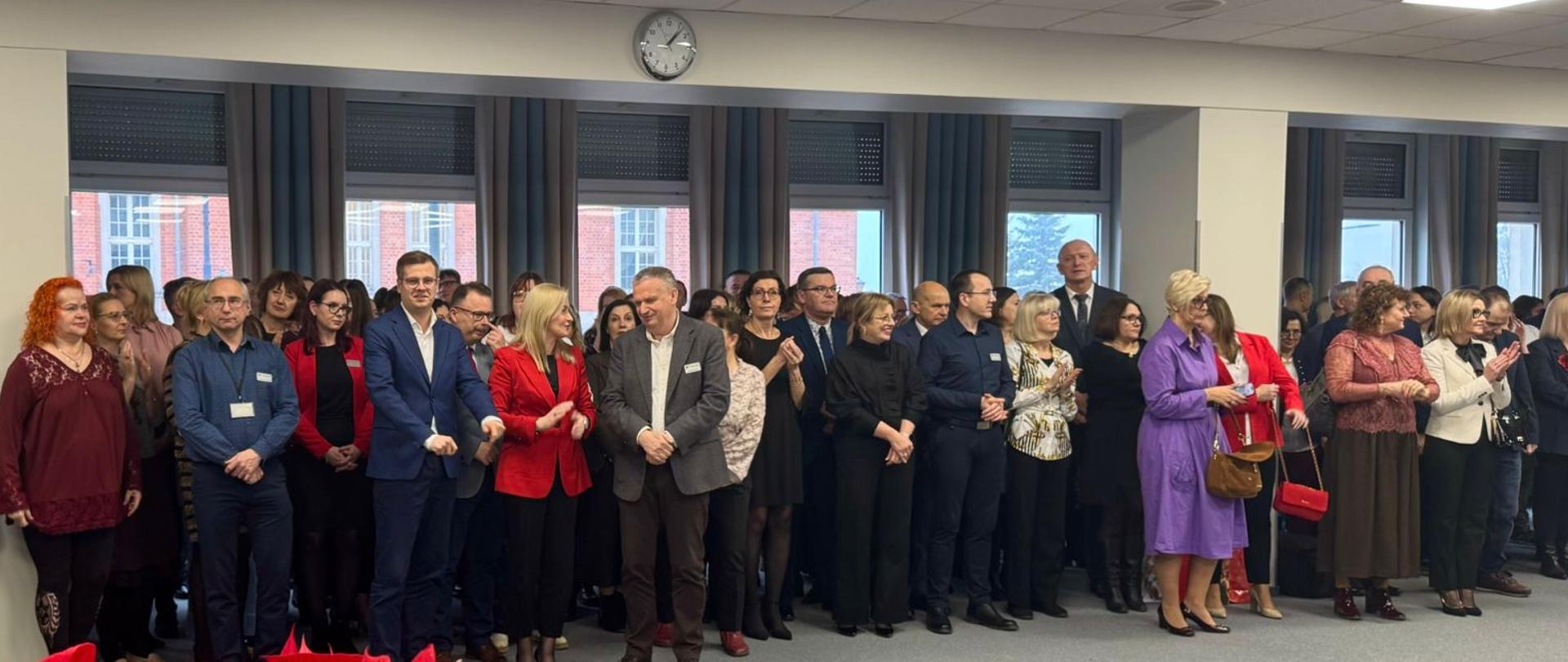 Duża grupa osób pozuje do zdjęcia w świątecznie udekorowanej sali, za plecami okna przez które widać czerwony budynek