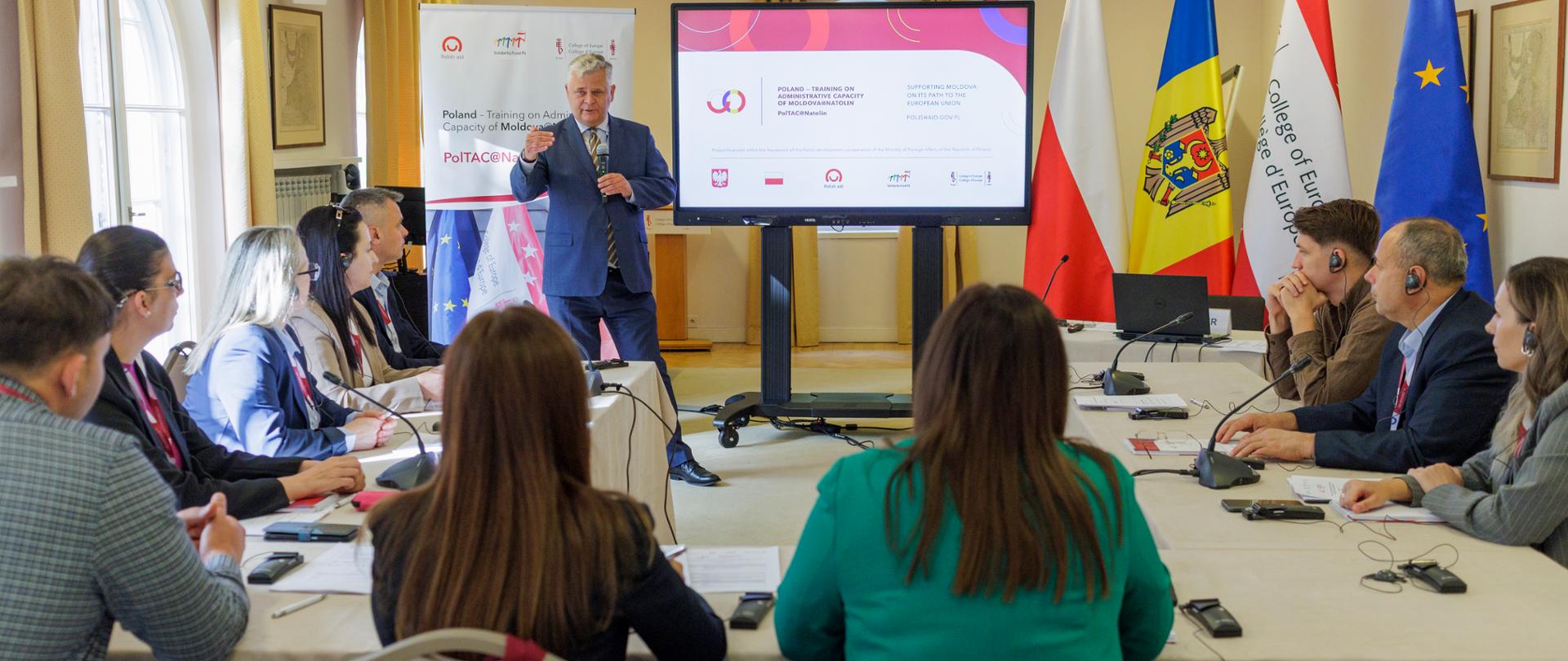 Przy stole konferencyjnym siedzi grupa osób i słucha mężczyzny prowadzącego szkolenie 