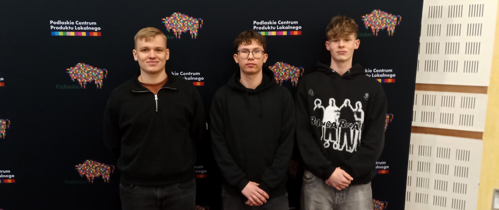 Trzej młodzi chłopcy stoją na tle ciemnej ścianki wystawienniczej z powtarzającymi się logotypami Podlaskiego Centrum Produktu Lokalnego.