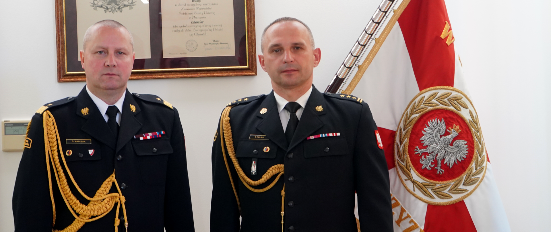 Na zdjęciu widzimy dwóch mężczyzn strażaków pozujących do zdjęcia. strażacy ubrani są w galowe mundury koloru czarnego, złote sznury na prawym ramieniu. W tle widzimy dyplom w ramie wiszący na białej ścianie. Z prawej strony kadru widzimy część sztandary koloru biało-czerwonego, w środku biały orzeł ze złota koroną. 
