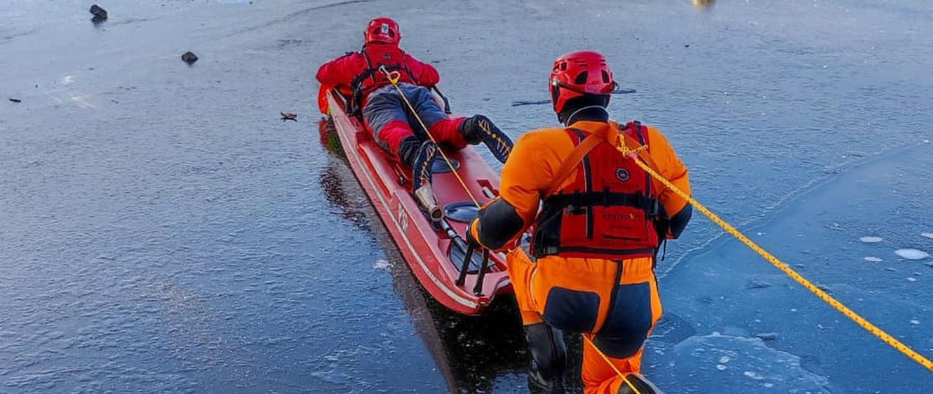 Strażacy PSP w zabezpieczeniu w kamizelkach ratowniczych oraz ubraniach wypornościowych podczas ćwiczeń z wykorzystaniem sań lodowych. Mundurowi asekurowani linami podążają do manekina umieszczonego na lodzie. Jeden ze strażaków leży na saniach lodowych, drugi w pozycji klęczącej asekuruje pierwszego na końcu sań.