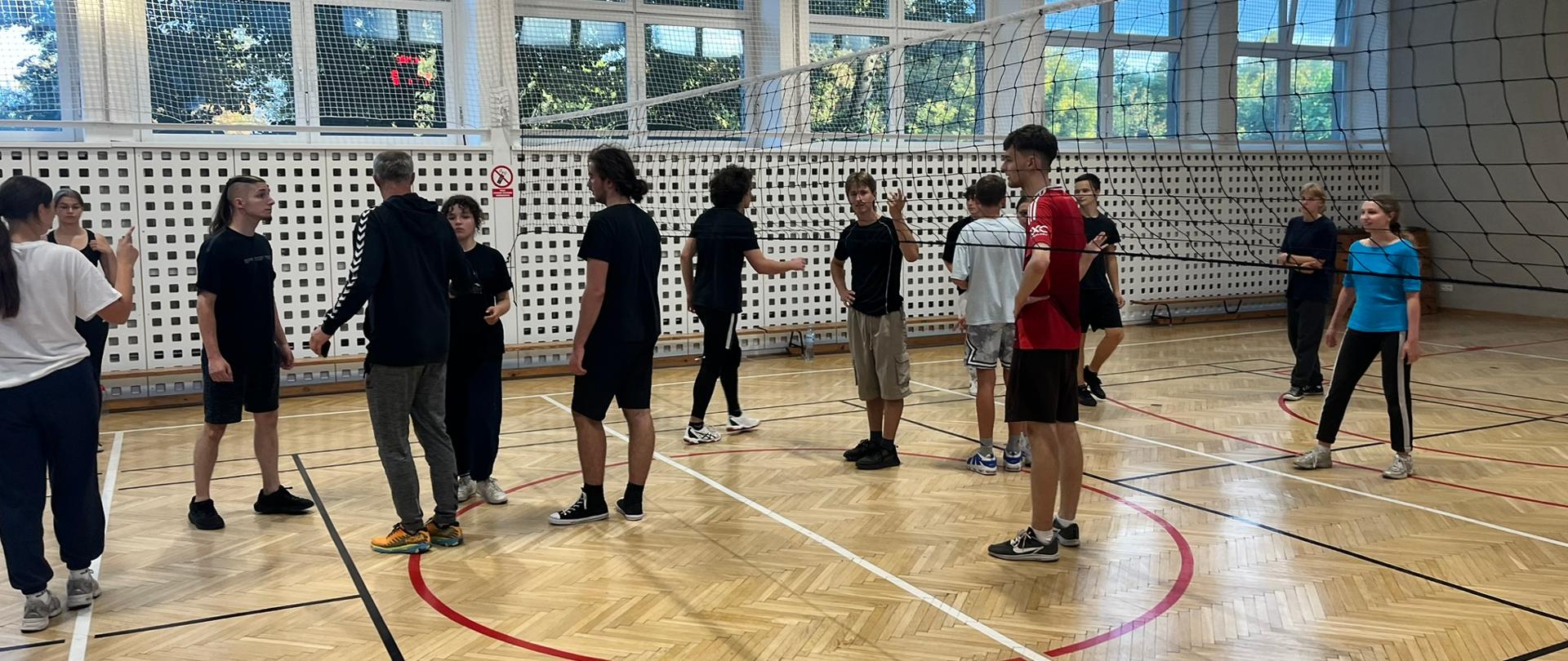 W sali gimnastycznej kilkunastoosobowa grupa dziewcząt i chłopców stoi po obydwu stronach siatki i przygotowuje się do gry w siatkówkę. Są ubrani w stroje sportowe.