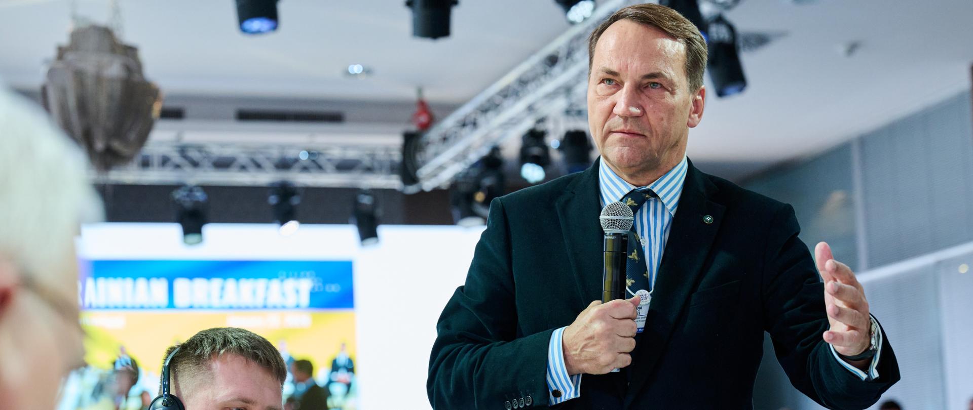 Mężczyzna w formalnym garniturze stoi i przemawia do publiczności, trzymając mikrofon w jednej ręce i gestykulując drugą. W tle widać salę konferencyjną z oświetleniem scenicznym oraz siedzących uczestników wydarzenia, w tym osobę w słuchawkach.