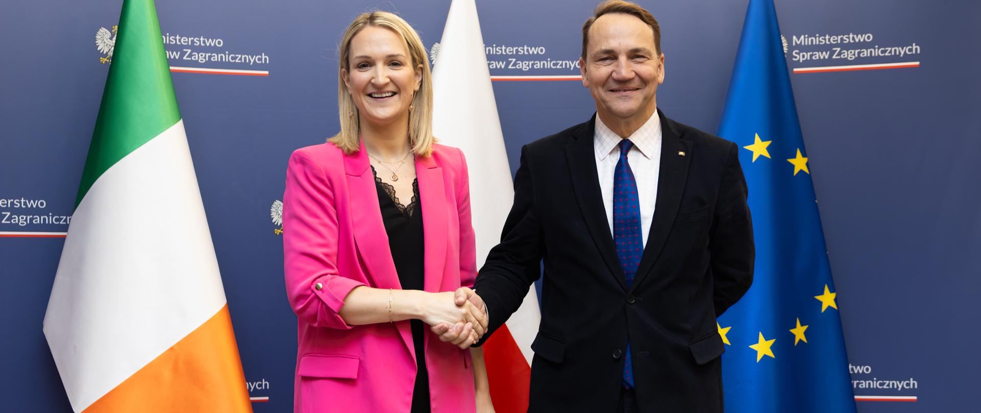 Na zdjęciu są kobieta i mężczyzna. Podają sobie dłonie. Patrzą w kierunku obiektywu. Za nimi jest ścianka z napisem "Ministerstwo Spraw Zagranicznych" i trzy flagi: Irlandii, Polski i Unii Europejskiej. 