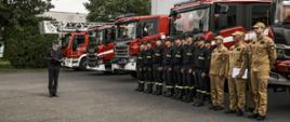 Dowódca zmiany odczytuje rozkaz dzienny, strażacy stoją w szyku na tle samochodów pożarniczych