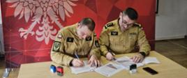 Dwóch funkcjonariuszy Państwowej Straży Pożarnej podpisuje dokumenty przy stole na tle ścianki Komendy Powiatowej PSP w Lwówku Śląskim.
