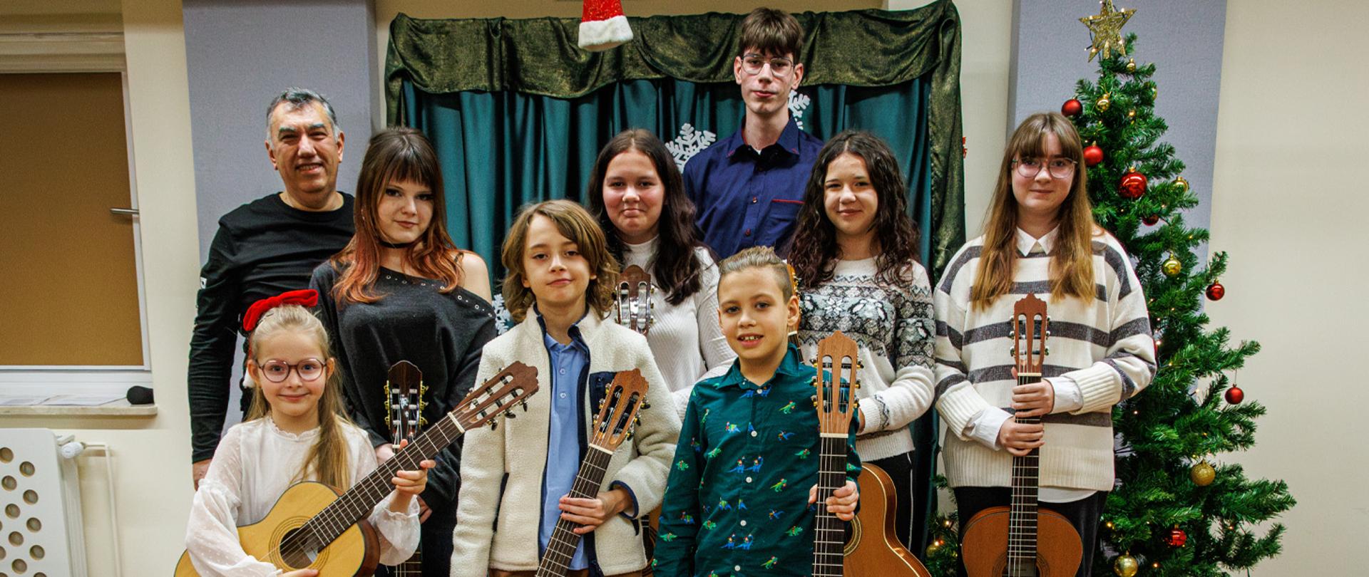 Nauczyciel z uczniami stoją w auli szkolnej, uczniowie trzymają w rękach gitary, za nimi stoi przystrojona choinka.