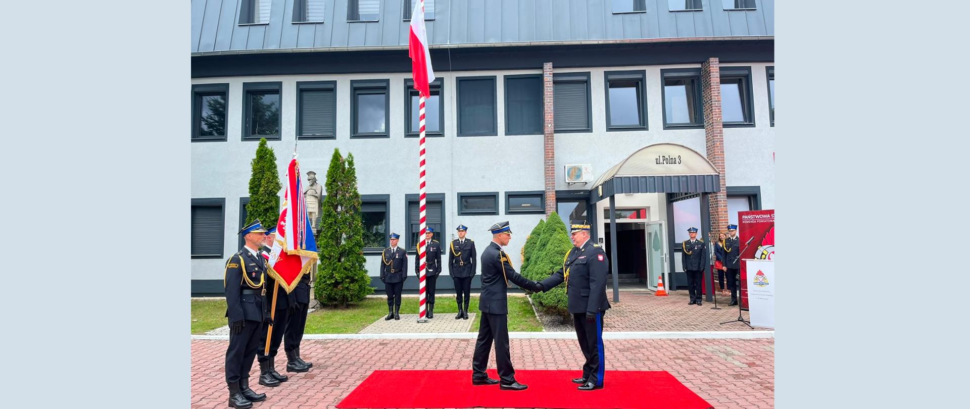 Plan pierwszy (główny punkt uwagi)
Uścisk dłoni: W centrum kadr, na rozłożonym prostokątnym czerwonym dywanie, stoi dwóch funkcjonariuszy Straży Pożarnej w galowych, ciemnogranatowych (lub czarnych) mundurach, czapkach rogatywkach oraz ze złotymi akselbantami (ozdobnymi sznurami) na ramionach.
Po lewej stronie młodszy strażak stoi profilem, podając dłoń starszemu rangą przełożonemu.
Po prawej stronie wyższy rangą dowódca w mundurze z oznaczeniami i polską flagą na ramieniu wita go uściskiem dłoni.
Plan drugi i tło
Poczet sztandarowy (po lewej stronie): Po lewej stronie dywanu stoi grupa strażaków trzymających uroczysty sztandar w barwach narodowych (biało-czerwono-niebieski ze złotymi godłami i frędzlami).
Warta / Szereg (w tle): W głębi, baczność trzyma trzech strażaków ustawionych w równym rzędzie przed maszt flagowy.
Maszt flagowy: Przed budynkiem stoi wysoki maszt w biało-czerwone pasy, na którego szczycie powiewa flaga Polski.
Kolejni strażacy i i goście (po prawej stronie): Przy wejściu do budynku oraz obok mównicy z logo Państwowej Straży Pożarnej stoją inni mundurowi oraz zaproszeni goście w strojach cywilnych.