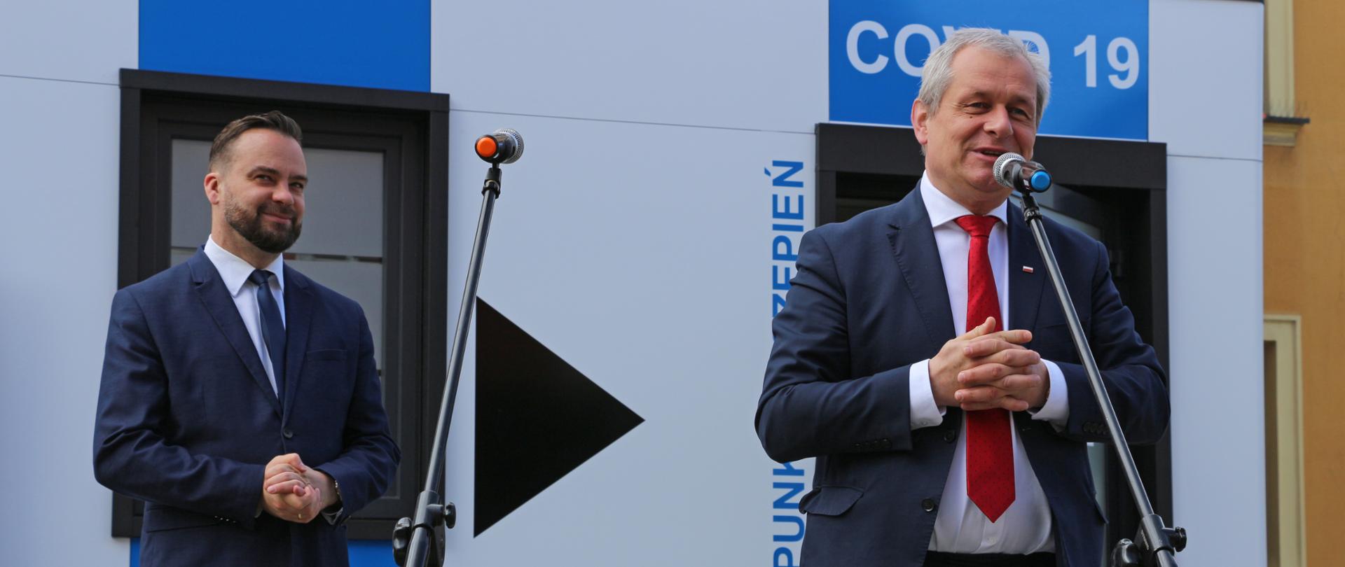 Na zdjęciu po prawej stronie kadru wojewoda opolski, stojący za mikrofonem - przemawia do dziennikarzy. Wojewoda patrzy w dal, ma splecione dłonie. Obok wojewody, po jego prawej stronie, stoi burmistrz Namysłowa. Mężczyzna stoi nieco w oddali, spogląda w dal. Stoi w lekkim rozkroku. W tle, za mężczyznami kontener szczepienny. Kontener zawiera napisy Covid-19 oraz mobilny punkt szczepienny. Kontener jest duży, prostokątny. Na zdjęciu widać jedynie jego front. Po prawej stronie kadru, przed kontenerem, widać fragment stołu, okryty białym obrusem. 
