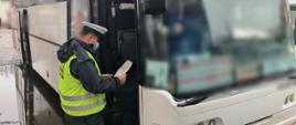 Inspektor stoi przed otwartymi drzwiami kontrolowanego autobusu. W ręku trzyma dokumenty. 