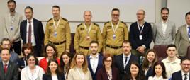 Grupowe zdjęcie uczestników wydarzenia, wykonane w przestrzeni konferencyjnej. Osoby ustawione są w trzech rzędach, uśmiechają się lub patrzą poważnie w stronę aparatu. Wśród nich znajdują się mężczyźni w piaskowych mundurach Państwowej Straży Pożarnej, a także kobiety i mężczyźni w strojach formalnych, z identyfikatorami na białych smyczach. Tło stanowi jasna ściana z ekranem, na którym częściowo widoczna jest prezentacja. 
