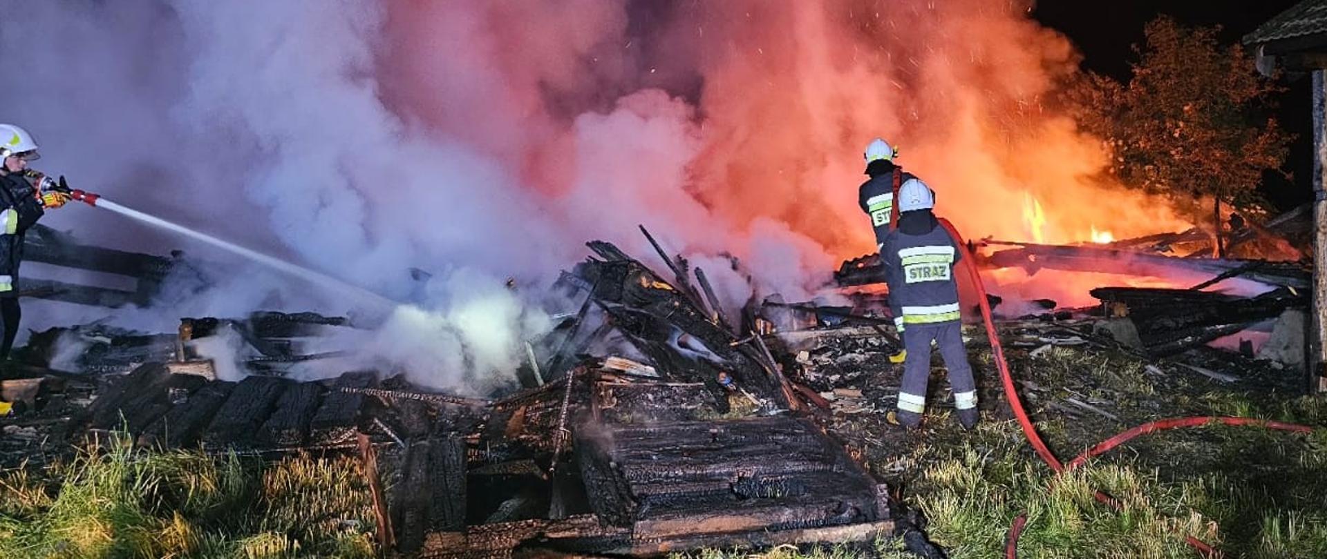 Zdjęcie przedstawia trzech ratowników walczących z pożarem. Centralnie widać zwęglone drewno stanowiące konstrukcję stodoły, głębiej: płomienie, dym oraz parę wodną. 
