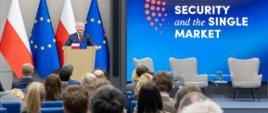 Widok sali z uczestnikami siedzącymi w rzędach, słuchającymi osoby mówiącej z mównicy na tle ekranu „Security and the Single Market”.