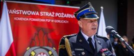 Na zdjęciu widoczny zastępca małopolskiego komendanta wojewódzkiego przemawiający podczas obchodów powiatowych dnia strażaka w Gorlicach. W tle widoczny rolap a na czerwonym tle napis koloru białego Państwowa Straż Pożarna Komenda Powiatowa Państwowej Straży Pożarnej w Gorlicach a obok flagi biało czerwone.