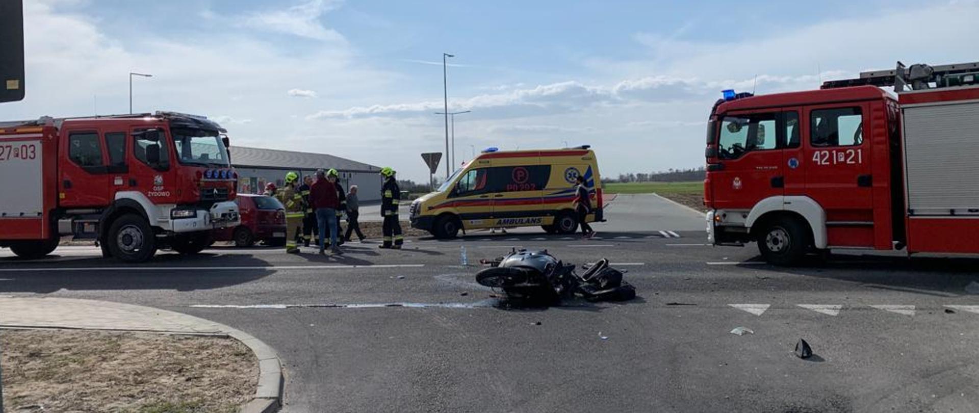 Słonecznie. Widok z boku drogi. Na środku asfaltowej jezdni leży ciemny rozbity motocykl. Za nim żółta karetka Pogotowia Ratunkowego. Po obu stronach czerwone pojazdy Straży Pożarnej. Na jezdni stoją strażacy w jasnych i ciemnych mundurach.