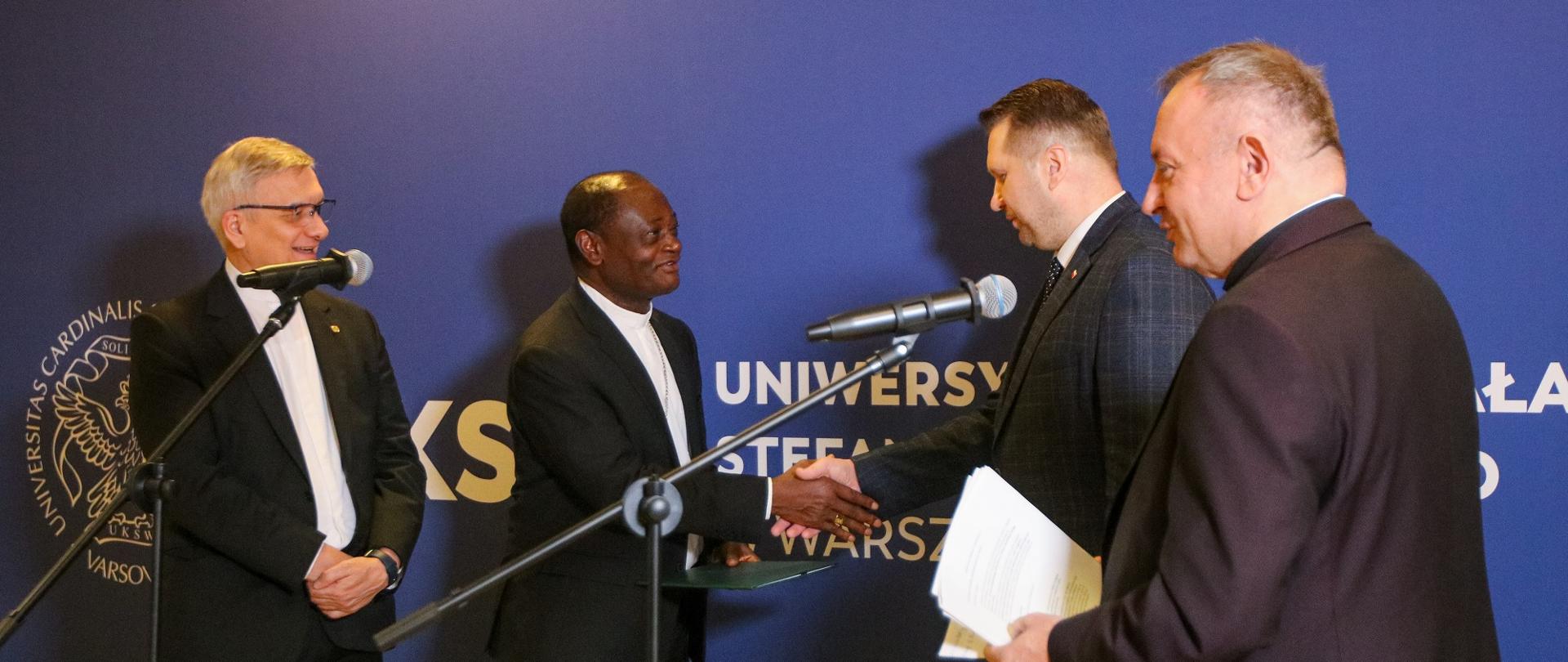 Minister Czarnek podaje rękę czarnoskóremu mężczyźnie w ciemnej marynarce, obok nich stoi dwóch mężczyzn, za nimi niebieska ściana w napisem UKSW.