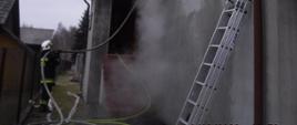 W głównej części zdjęcia widzimy dom mieszkalny 3-kondygnacyjny z okna którego wydostaję się dym oraz para wodna. Po lewej stronie widzimy strażaka podtrzymującego linię wężową, która skierowana jest do okna na piętrze. Po prawej stronie widać drabinę opartą o budynek. W oddali, po lewej stronie widać dachy sąsiednich budynków. U dołu zdjęcia widzimy dużą ilość odcinków wężowych, które mają na celu dostarczanie wody.