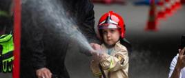Na zdjęciu widać chłopca w hełmie oraz ubiorze strażackim, który leje wodę do celu. Porad udziela mu asystujący obok mu strażak.