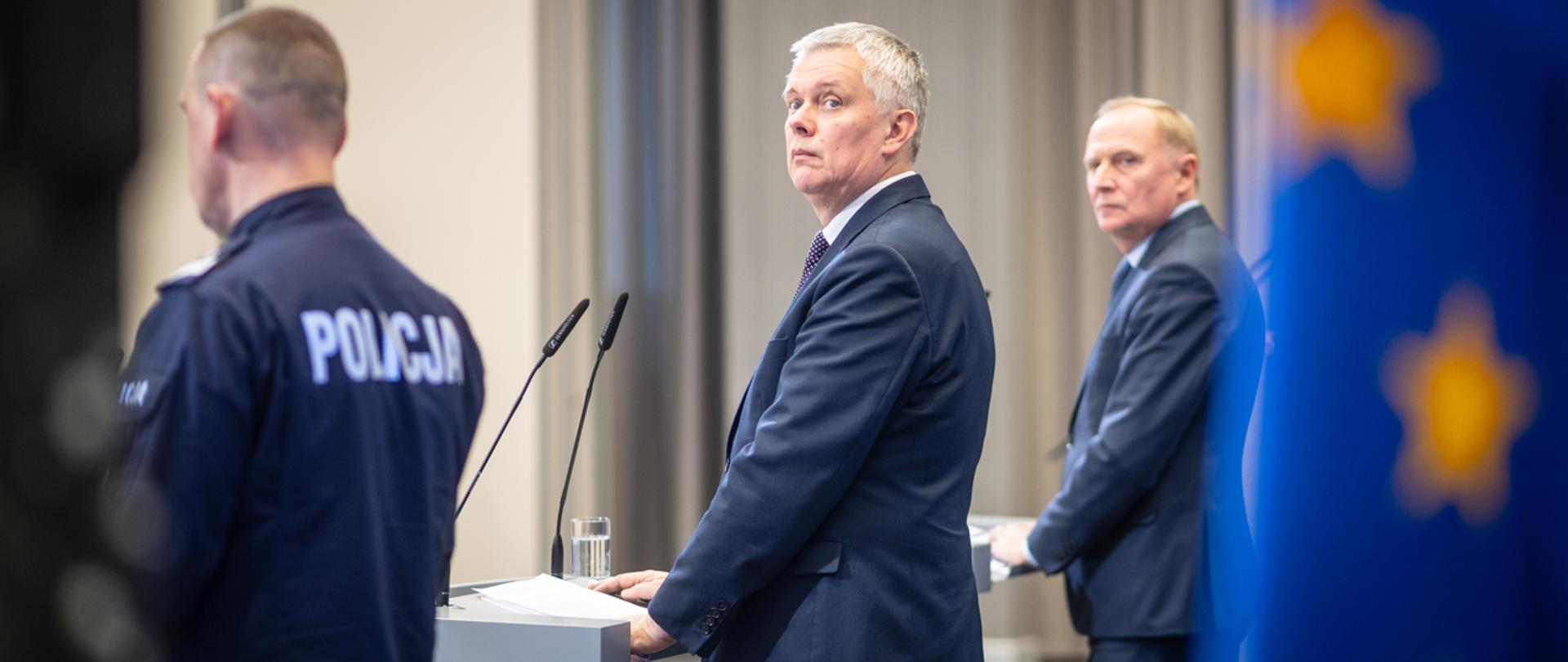 Trzech mężczyzn stojących tyłem, z których ten który stoi na środku jest zwrócony za siebie. Po prawej w nieostrości flaga UE. 