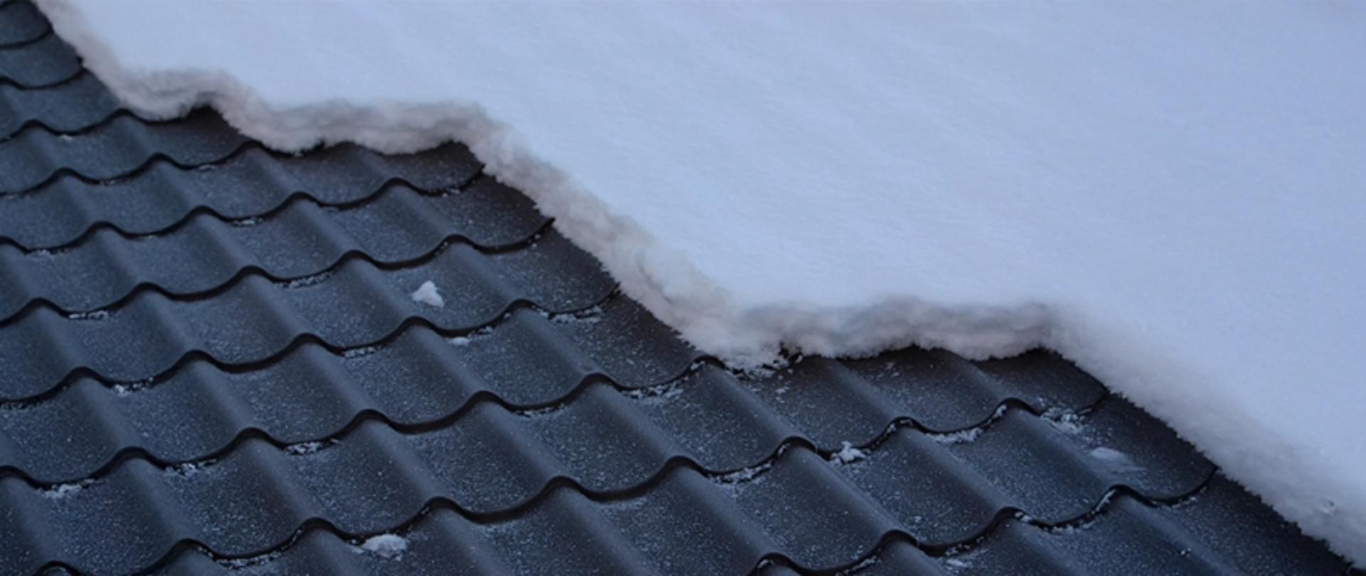 Dach z ciemnej dachówki falistej, górna część pokryta warstwą śniegu, dolna odsłonięta.
