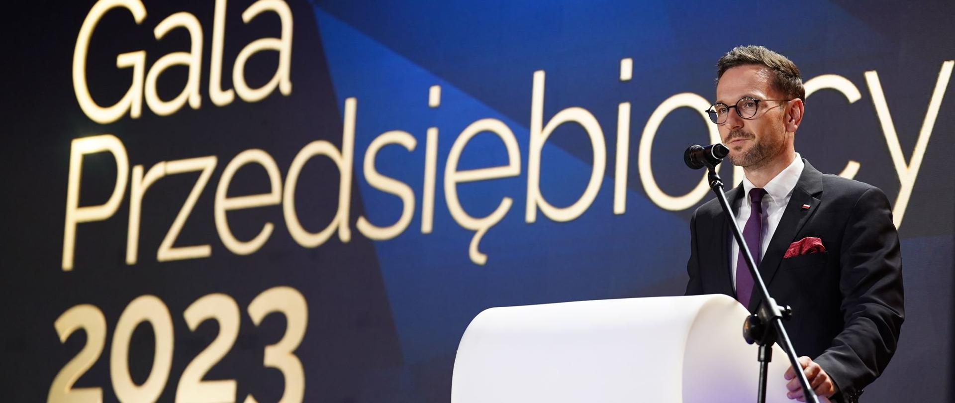 Gala Przedsiębiorcy 2023 w MRiT. Minister Waldemar Buda stoi po prawej stronie przy białej mównicy. W tle napis: Gala Przedsiębiorcy 2023.