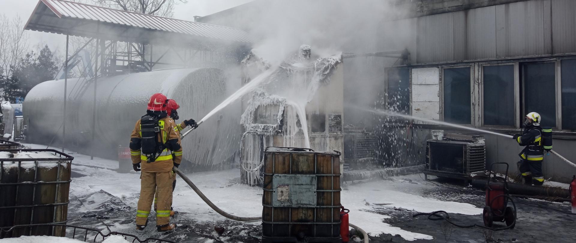 Zdjęcie przedstawia trzech strażaków gaszących chłodnicę na terenie zakładu przetwarzającego gumę. Z chłodnicy wydobywa się dym. Po lewej i po prawej stronie chłodnicy o kształcie dużego kwadratu stoją strażacy i polewają chłodnicę pianą. Strażacy ubrani są w aparaty ochrony układu oddechowego.
