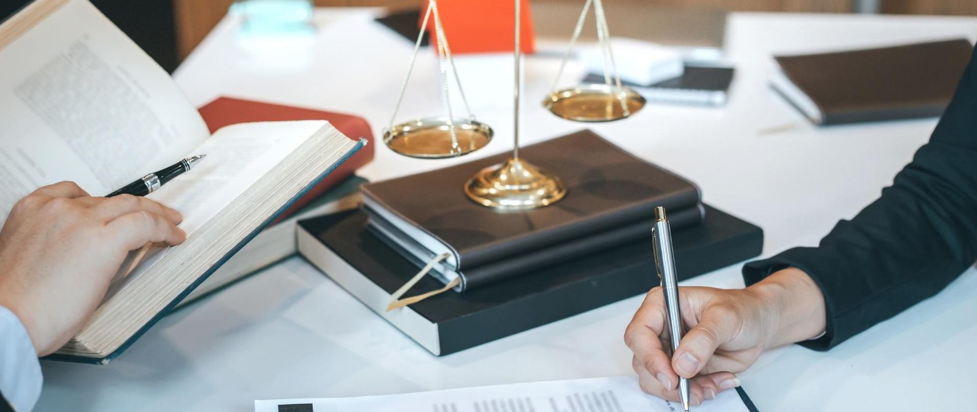 Grafika, ktoś pisze długopisem na kartce, obok ktoś inny trzyma otwartą książkę, między nimi stoi mała szalkowa waga.