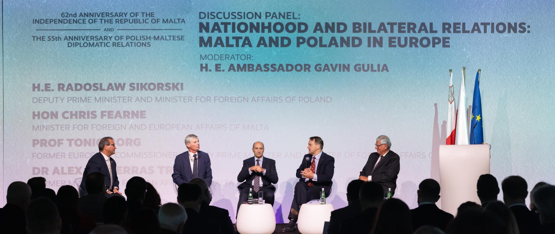 The photo shows five men in suits sitting on stage and speaking to the audience. Behind them is a screen with the words “discussion panel: nationhood and bilateral relations: Malta and Poland in Europe”