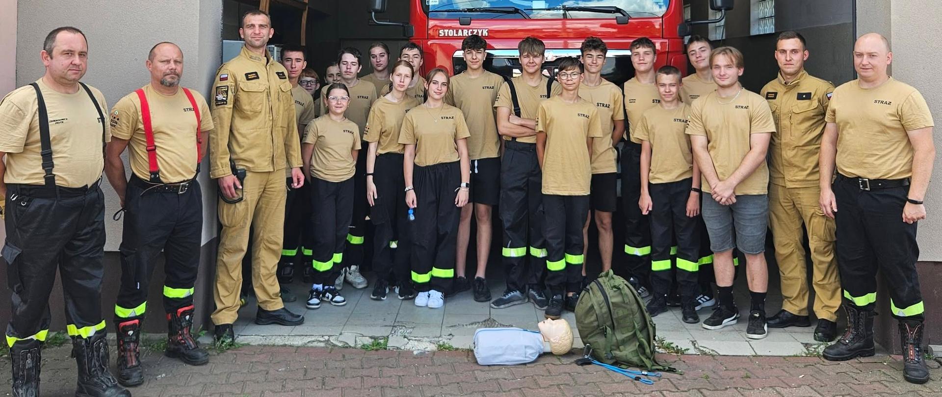 Na zdjęciu grupa młodzieży oraz strażaków pozuje do wspólnej fotografii przed garażem jednostki straży pożarnej. W tle znajduje się samochód ratowniczo-gaśniczy. Uczestnicy spotkania mają na sobie odzież strażacką. Na pierwszym planie widoczny jest sprzęt wykorzystywany do udzielania pierwszej pomocy, w tym plecak ratowniczy i fantom do nauki resuscytacji krążeniowo-oddechowej.