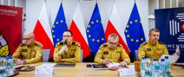 Czterech oficerów PSP podczas konferencji prasowej. Na środku głos zabiera Komendant Główny. W tle cztery flagi Polski i trzy flagi Unii Europejskiej, a nad nimi godło Rzeczypospolitej Polskiej.