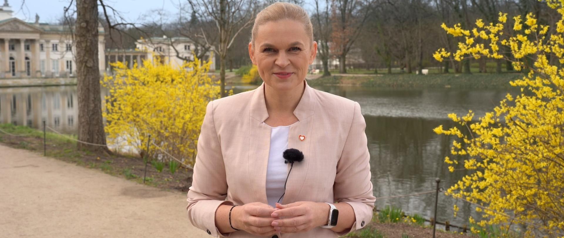 Minister Barbara Nowacka składa życzenia wielkanocne. w tle wiosenne żółte krzewy. Życzenia świąteczne minister Barbary Nowackiej. „Zaczynamy wiosnę w polskiej edukacji”