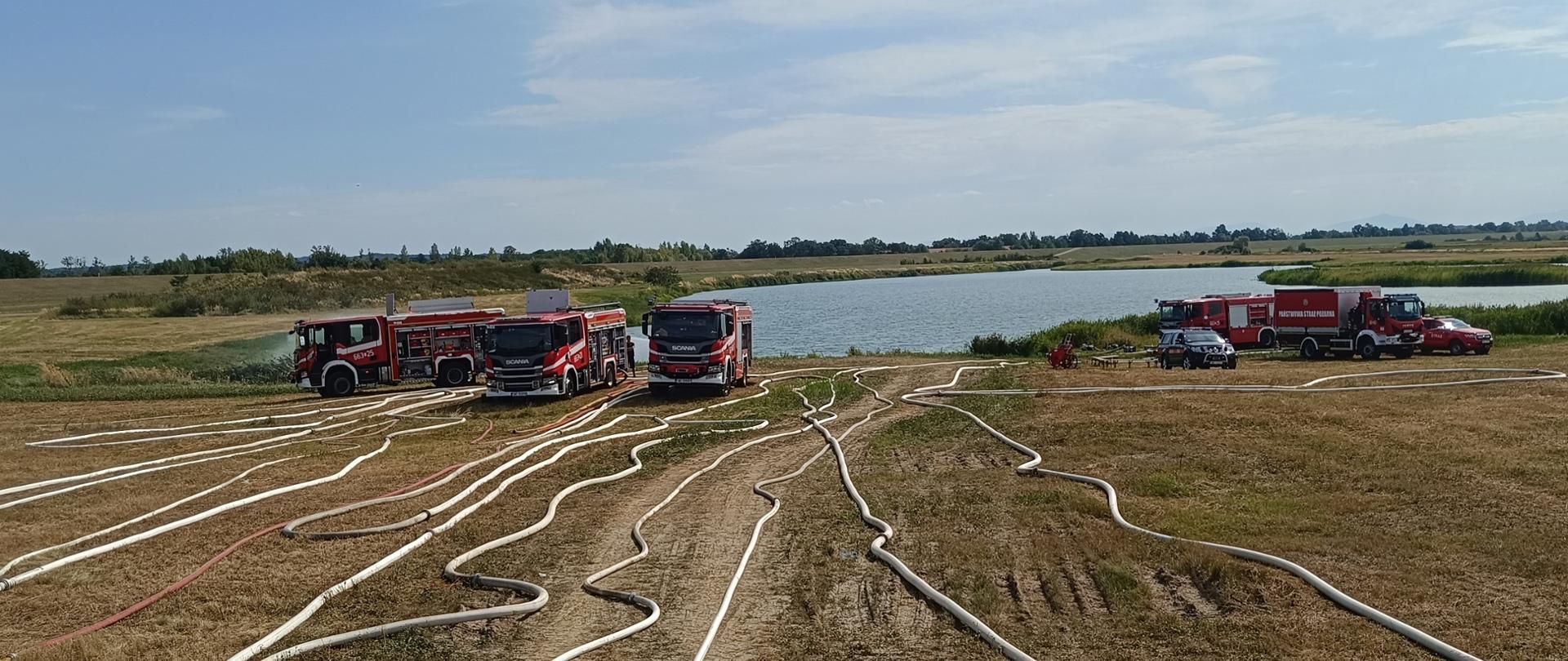Trzy wozy strażackie podczas pompowania wody do widocznych węży znajdujących się przed nimi. W tle naturalny zbiornik wodny.