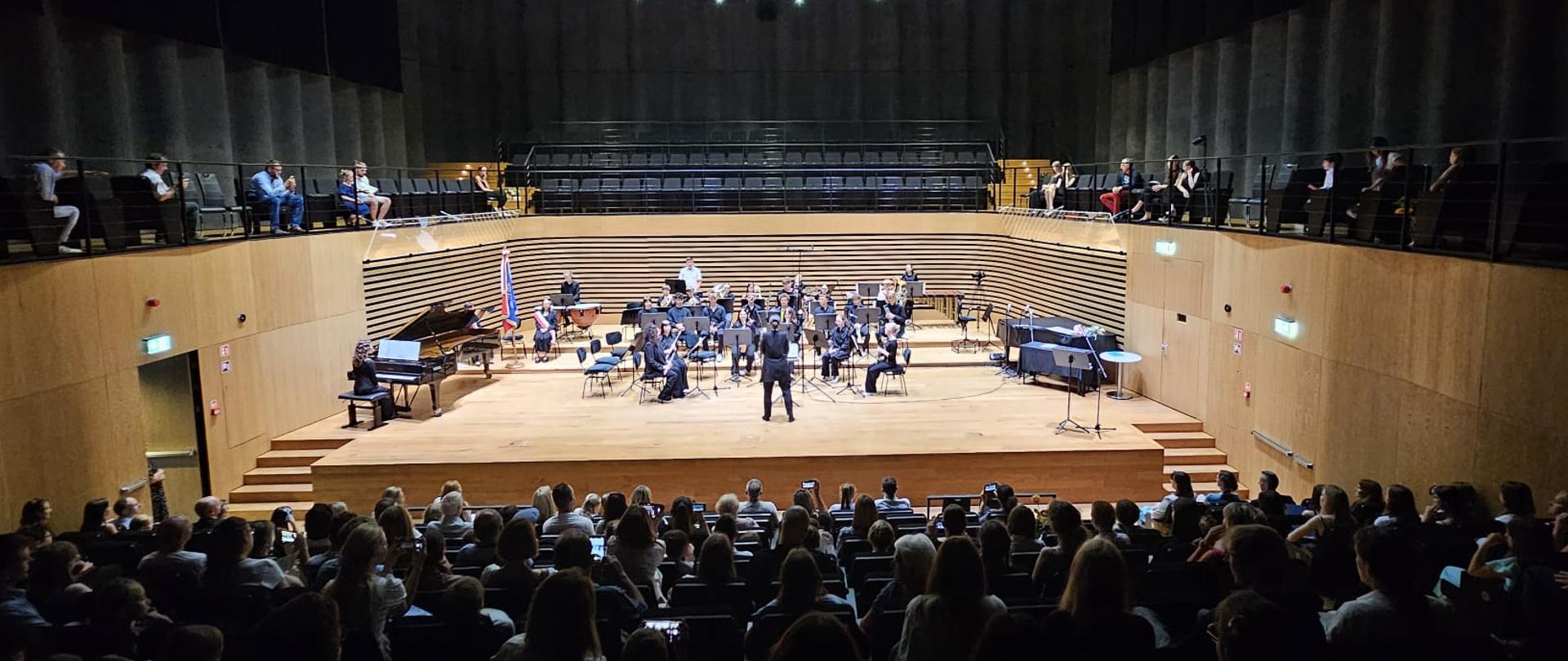 Kobieta dyryguje orkiestrą, z przodu widoczna od tyłu widownia zapełniona publicznością.
