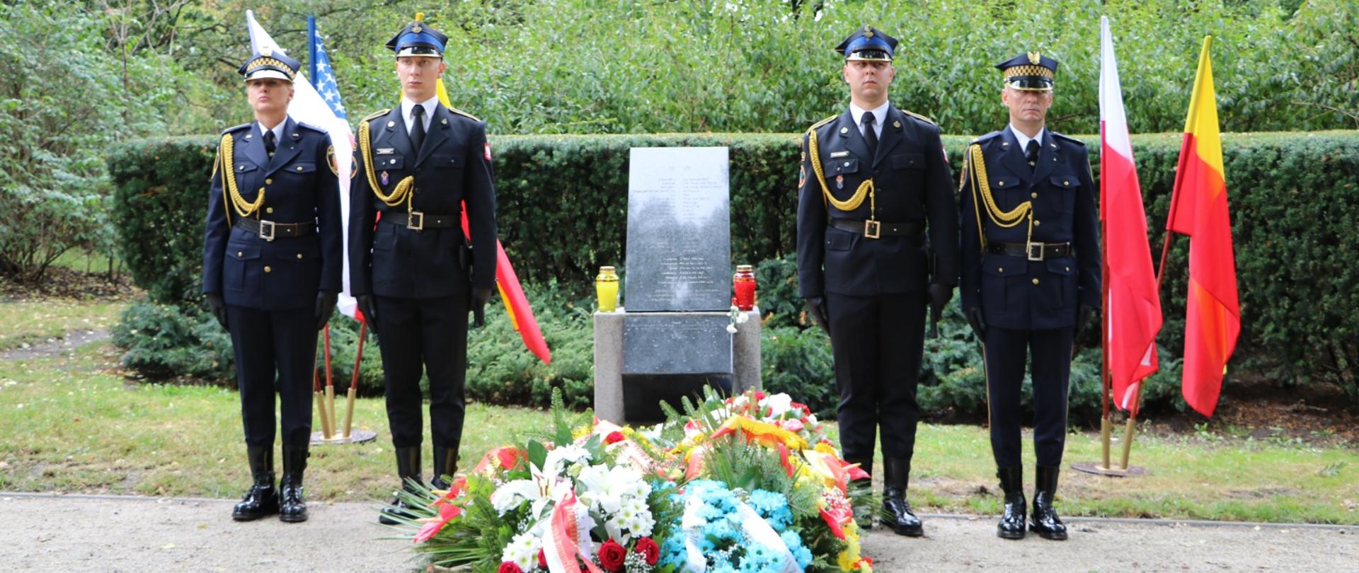 Zdjęcie przedstawia uroczystą ceremonię. Cztery osoby w granatowych mundurach ze złotymi akcentami, czapkach i czarnych butach stoją przed pomnikiem. Po obu stronach pomnika widać dwie flagi; po lewej znajduje się flaga Stanów Zjednoczonych, a po prawej – czerwono-żółta flaga, obok czerwono-biała flaga, która jest flagą Polski.
U podstawy pomnika leży duży, kolorowy wieniec z kwiatów. Wieniec, umieszczony na żwirowej ścieżce, składa się z różnych kwiatów, w tym białych, czerwonych i jasnoniebieskich, a także zielonych liści i biało-czerwonych wstążek. Na podstawie pomnika znajduje się również świeca w czerwonym szklanym pojemniku oraz inna świeca lub przedmiot w żółtej obudowie. Tło stanowią drzewa i zielone krzewy. Niebo jest zachmurzone. 