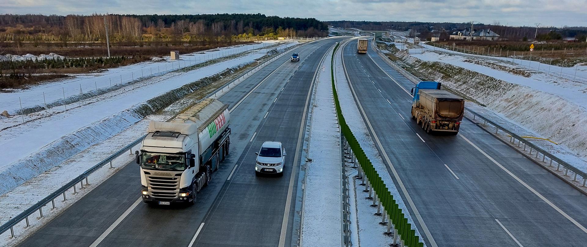 Dwujezdniowa autostrada z pasem rozdzielającym, na którym zamontowano zielone bariery energochłonne. Droga jest sucha, ale pobocza i otoczenie są pokryte cienką warstwą śniegu.
Na lewej jezdni (kierunek w stronę obserwatora) jedzie duży biały ciągnik siodłowy z naczepą oraz samochód osobowy. Na prawej jezdni (kierunek od obserwatora) widoczna jest ciężarówka z odkrytą skrzynią ładunkową.
W oddali widać kolejne pojazdy, głównie ciężarówki. Po obu stronach autostrady rozciągają się pola i zadrzewienia, a na horyzoncie widoczny jest las. Niebo jest częściowo zachmurzone, z przebłyskami błękitu.
Dominują odcienie szarości asfaltu, bieli śniegu oraz zieleni barier. Niebo ma mieszankę jasnych chmur i niebieskich fragmentów.