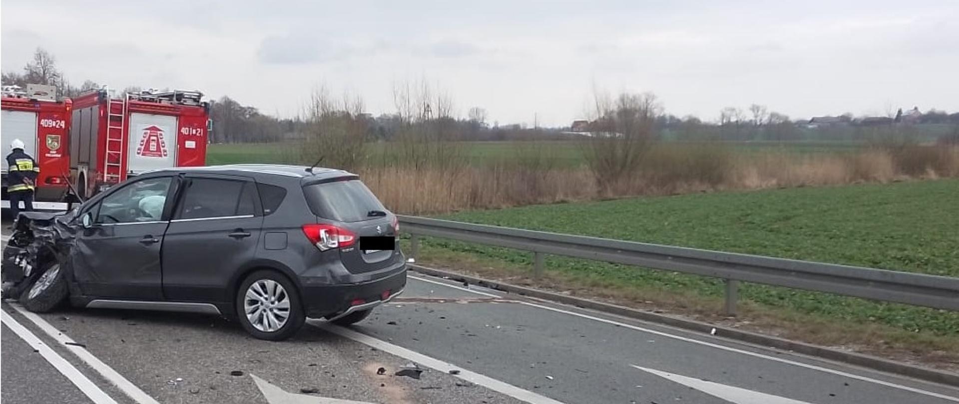 Samochód osobowy z uszkodzonym przodem stoi na asfalcie, w tle samochody strażackie, na prawo widać tereny zielone
