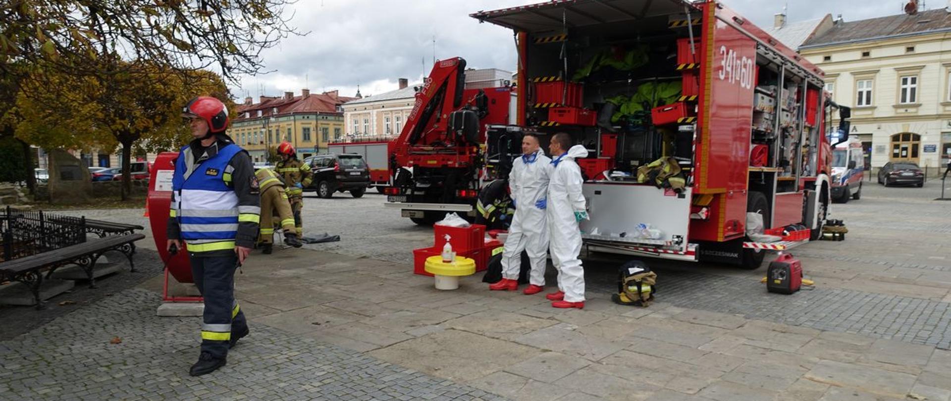 Strażacy kombinezonach oraz samochody straży pożarnej.