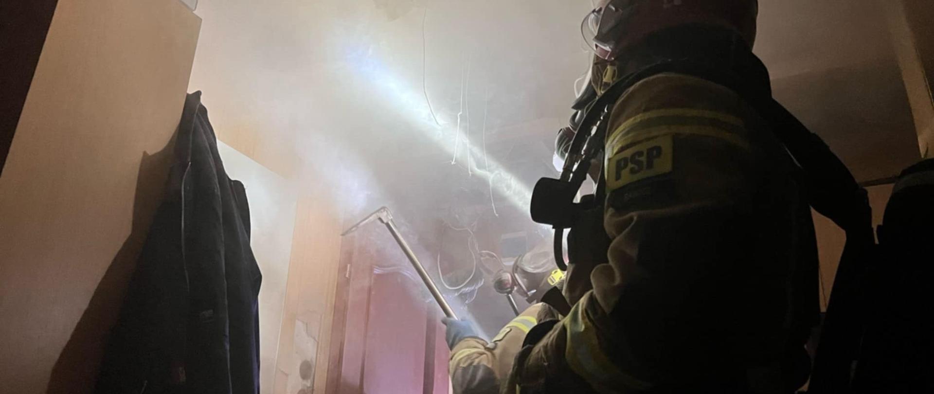 Wnętrze mieszkania, w którym powstał pożar. Z sufitu zwisają przewody elektryczne. Występuje silne zadymienie. W pomieszczeniu jest 2 strażaków wyposażonych w aparaty ochrony układu oddechowego. Jeden z nich wykonuje prace rozbiórkowe przy pomocy hooligana.