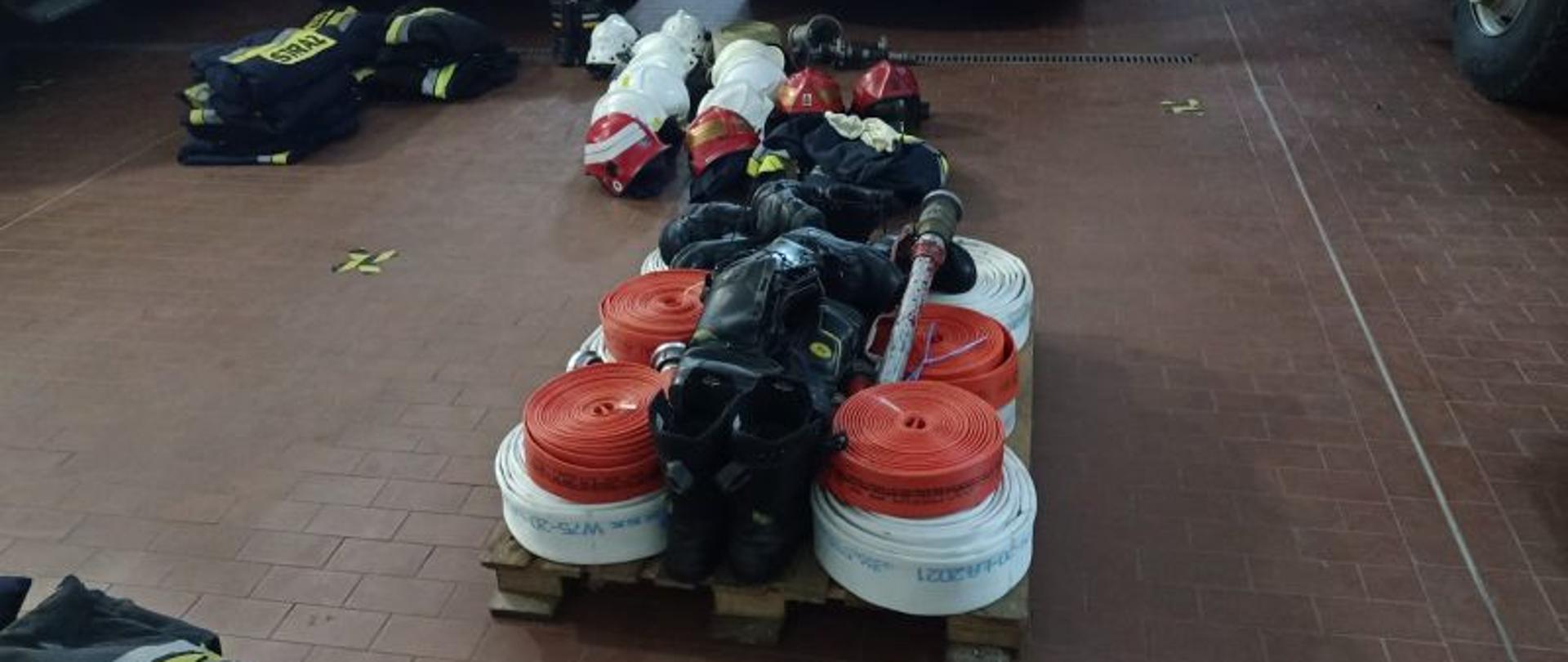 Centralny punkt zdjęcia stanowią przygotowane na palecie węże strażackie, buty, prądownicę. W tle inny sprzęt. Wszystko zmagazynowane w garażu